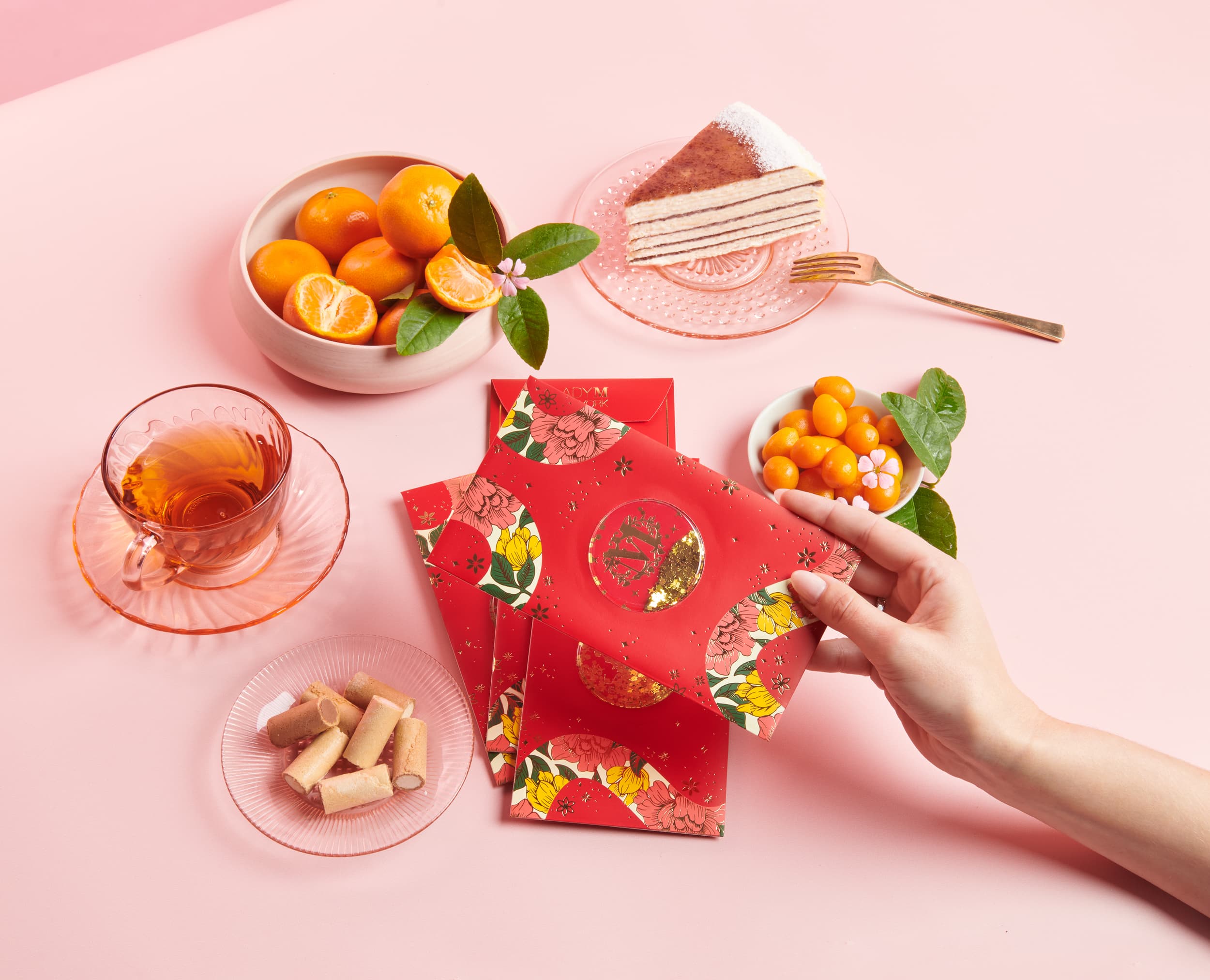 A festive Lunar New Year setting with red floral-patterned envelopes, a cup of tea, a slice of layered cake, fresh oranges, and traditional snacks, all arranged on a pastel pink background.