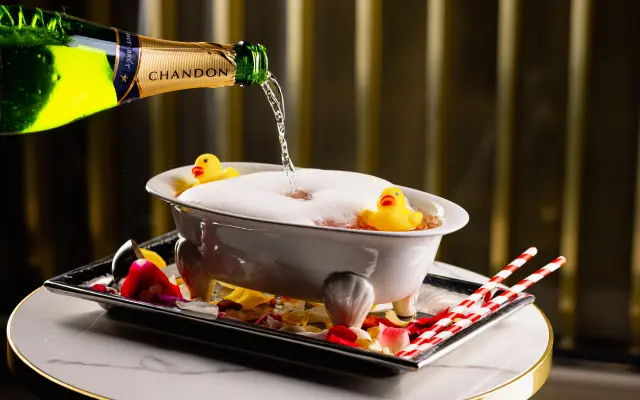 A bottle of Chandon champagne being poured into a small foam-filled bathtub with rubber ducks, placed on a tray with rose petals and straws.